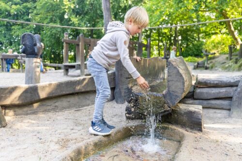 Kind hält Hand unter fließendes Wasser an der Mündung eins hohlen Baumstammes auf dem Wasserspielplatz